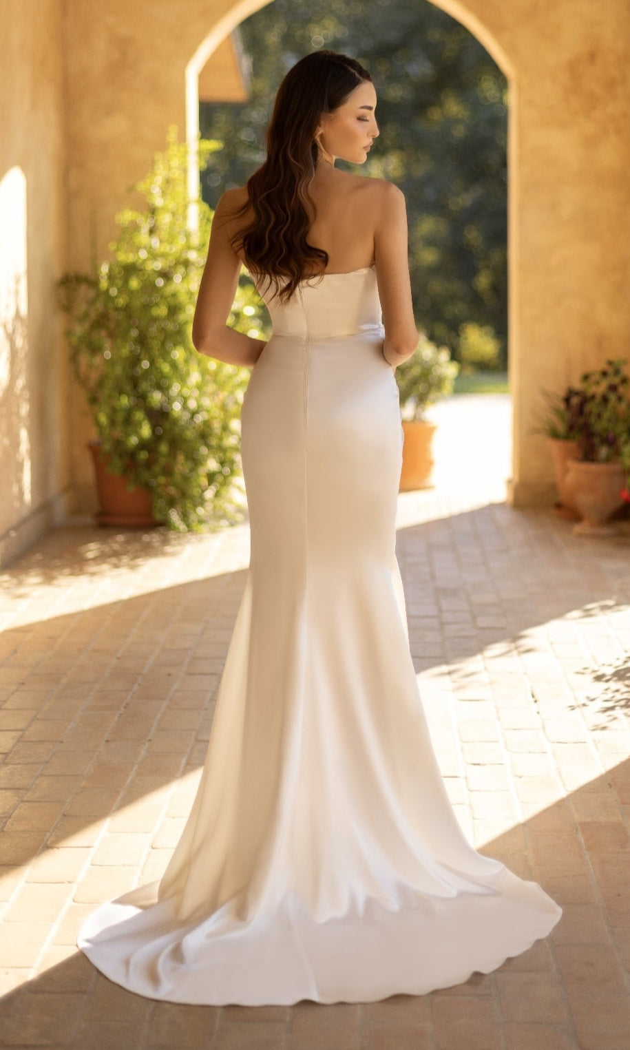 A woman in the Off-White Strapless Prom Gown: Tina Holly TT003W stands on a sunlit, tiled terrace with potted plants and an arched doorway in the background.